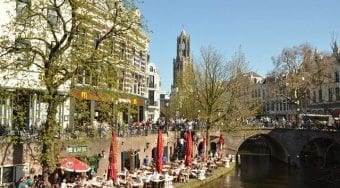 A restaurant with red umbrellas and lots of people set up along the banks of a canal in Amsterdam with trees and buildings surrounding it.