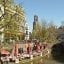 A restaurant with red umbrellas and lots of people set up along the banks of a canal in Amsterdam with trees and buildings surrounding it.