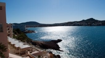 Panoramic view of the sparkling Mediterranean Sea along the rocky shoreline of Santa Ponsa, Mallorca, with sun reflecting off the water, terraced resort balconies in the foreground, and distant hills framing the bay under a clear afternoon sky—an inviting scene for travelers making the scenic journey from Palma Airport to Santa Ponsa.