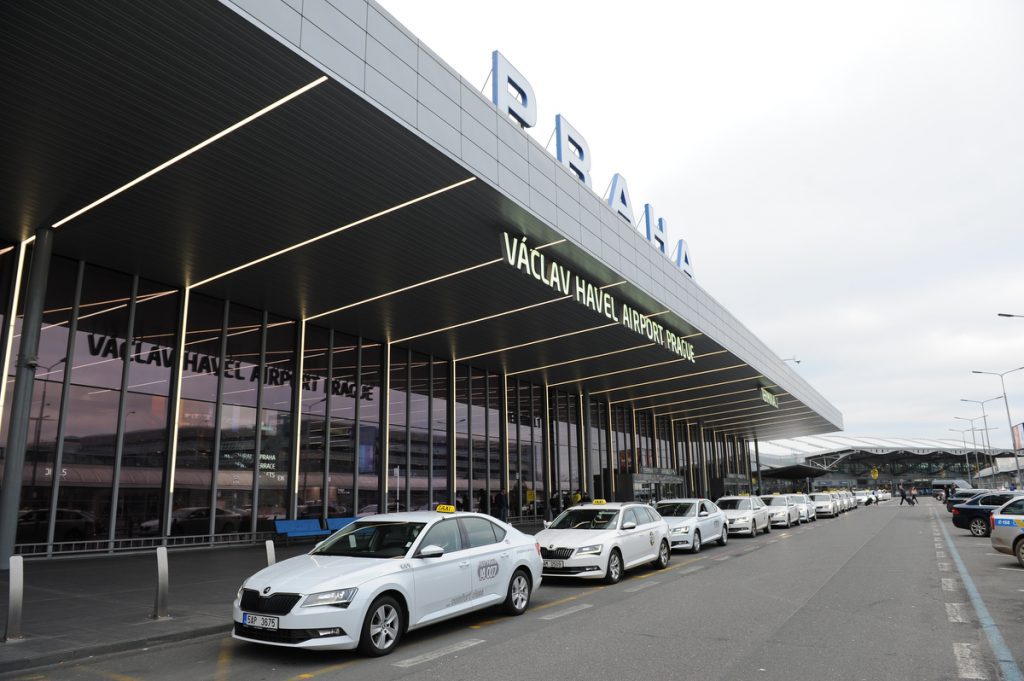 A wide view of white Prague Airport taxis lined up outside of the terminal, with PRAHA on the roof of the terminal.