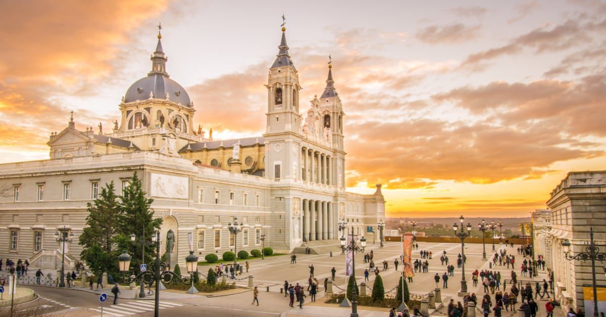 A huge ornate and white building in a large plaza filled with people and a setting sun in the background.