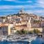 View of the Vieux Port in Marseille, France, with boats floating in the harbor and the Basilica of Notre-Dame de la Garde visible on top of a hill in the background.