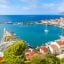 An enclosed bay with turquoise waters and a single sailboat surrounded by trees and white houses with terracotta roofs.