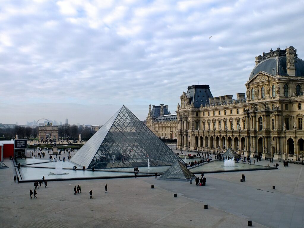 View of the Louvre Museum and glass pyramid in Paris on a cloudy afternoon, with visitors walking through the courtyard; an iconic stop during a layover in Paris.
