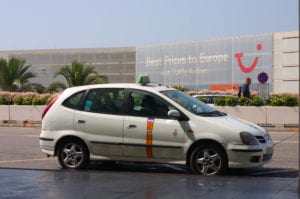 A white Mallorca taxi with a yellow stripe parked outside Palma de Mallorca Airport on a sunny day, ready to pick up arriving passengers.