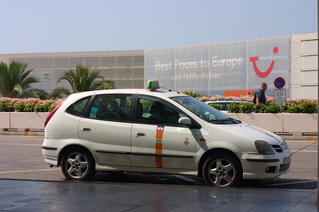 A white Mallorca taxi with a yellow stripe parked outside Palma de Mallorca Airport on a sunny day, ready to pick up arriving passengers.