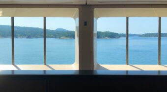 view from two ferry windows towards the outside, with blue water and green trees in the distance