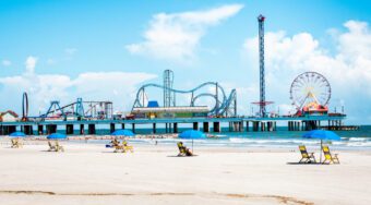 sandy beach with yellow sun-loungers. pier with amusement park in the background. Rides including rollercoasters and ferris wheels