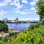 Scenic daytime view of downtown Ottawa across the river, framed by lush greenery and trees in the foreground. The skyline features modern buildings under a bright, partly cloudy sky.