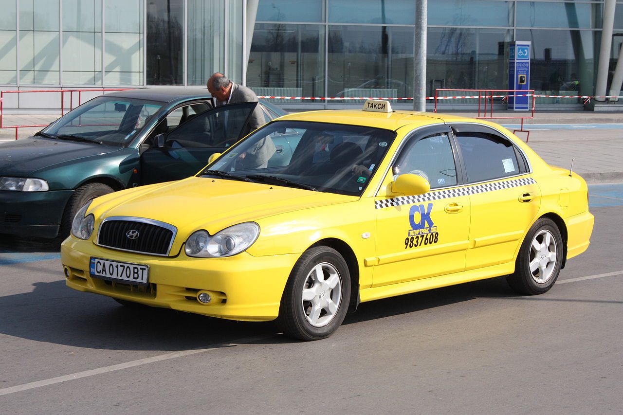 yellow taxi with company sign on the front door passing another car in front of the airport