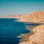 brown-beige mountains by the beach, dark blue waters under a baby blue sky at daytime