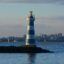 white and blue stripes on a lighthouse surrounded by body of water and a city appearing in the background