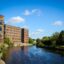 river flows by brown brick modern flats and an area full of green trees on the opposite side at Leeds Dock, Leeds, UK