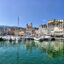 several small white boats in the green-blue waters by a deck near pale-paint coloured buildings under a clear sky