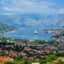 aerial shot of snowy mountain peaks at distance and a city by body of water, filled with green trees and red brick roofs in Kotor, Montenegro