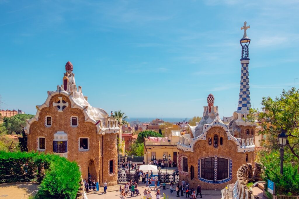 Barcelona's Park Guell entrance pavilions on a sunny day, made up of two Gaudi-style buildings and a blue and white checked tower with a cross on top.