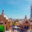 Barcelona's Park Guell entrance pavilions on a sunny day, made up of two Gaudi-style buildings and a blue and white checked tower with a cross on top.