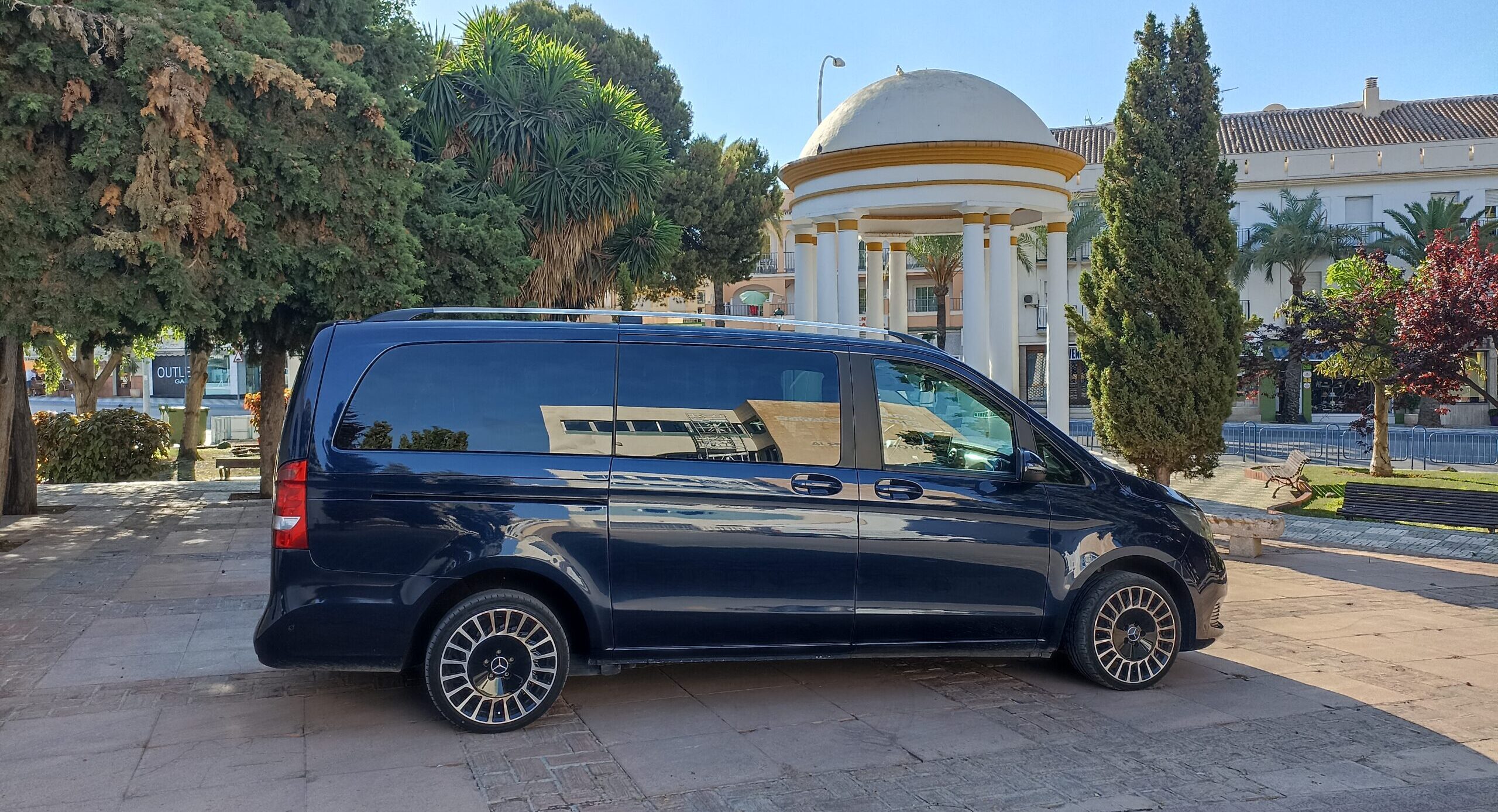 Faro Airport Taxi (Minivan) Side view of a black Faro Airport Taxi (Minivan) parked into front of trees and some buildings, with blue skies above.