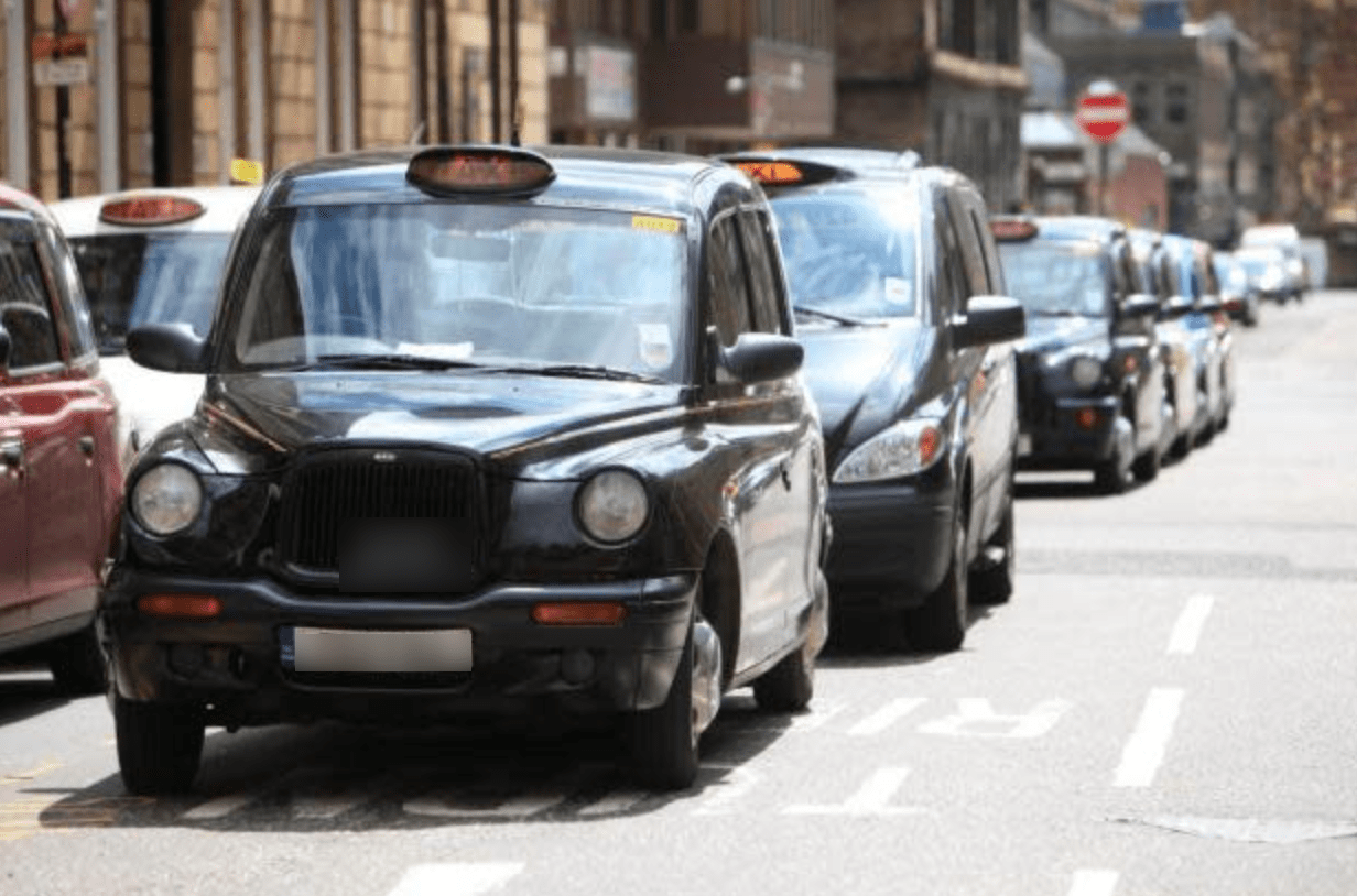 A black Hackney Glasgow cab at the front of a taxi rank, with a line of taxis parked behind it and another line to the left.