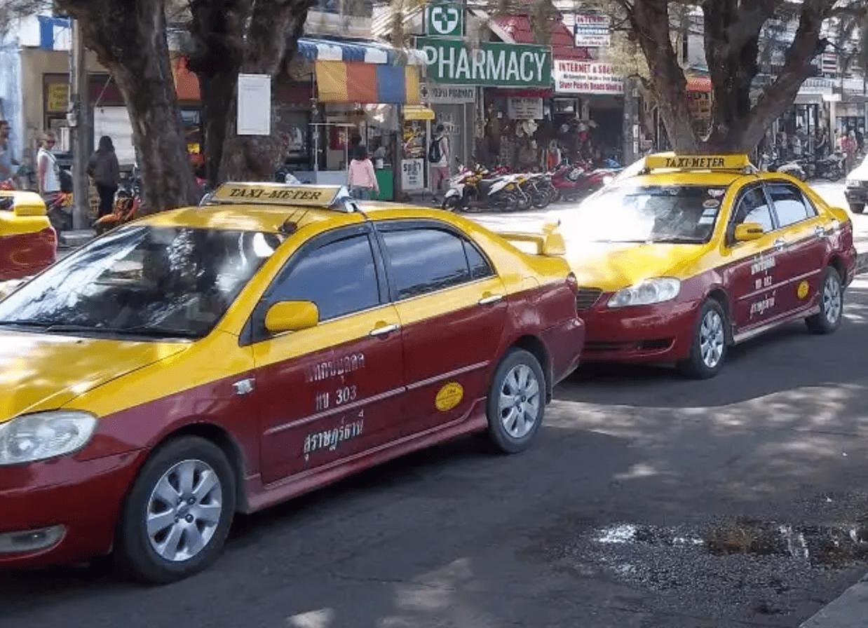 Koh Samui Taxi - Welcome Pickups Yellow and burgundy Koh Samui taxis lined up in a rank on the street, with shops behind.