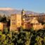 A view of the Alhambra perched on top of a hill surrounded by greenery, with mountains behind and blue skies above.