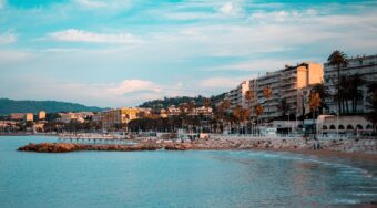 A sandy beach with blue waters and apartment buildings behind.