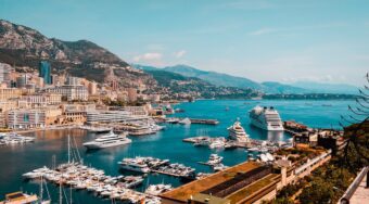 A view of Monaco's port with yachts in the marina and a view of the city and mountains behind.
