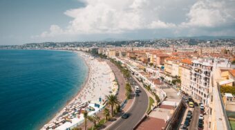 View of a palm-lined beach and the sea in Nice with a road and the city behind it and grey clouds above.