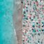 Birds eye view of a sandy beach filled with people and colourful sun umbrellas, and with holidaymakers swimming in turquoise blue waters.