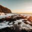 Aerial view of rolling waves hitting rocks at shore with a cliff in the background against the sun