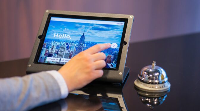 A hand clicking on a self-service touch screen with a hotel bell next to it.