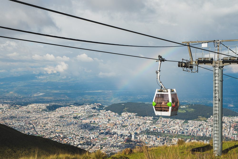 Panoramic view of Quito with a cable car hanging above it and a cloudy sky above with a rainbow. Quito Airport Photos