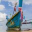 An iconic wooden Thai fishing boat on the beach, painted a blue colour with a yellow rim and red ribbons tied to it.