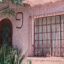 Front of a pink-walled house with a rounded front door and a window to the right, and plants growing in front.