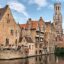 A row of old brick houses with terracotta roofs built on a riverbank with a church in the background