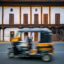 A yellow auto-rickshaw traveling at a high speed in front of a white building with wooden doors.
