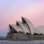 The Sydney Opera House set against a pink and blue sunset sky with the harbour in front.