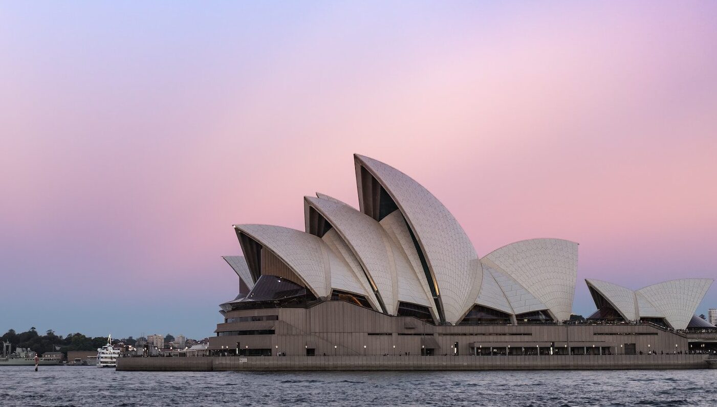 The Sydney Opera House set against a pink and blue sunset sky with the harbour in front.