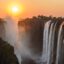 View of Victoria Falls waterfall at sunset, with an orange sky and sun above.