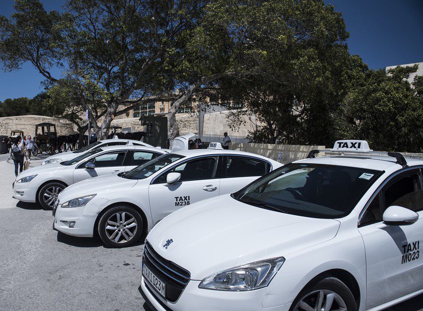 A line of white Malta taxis parked under the shade of a tree.
