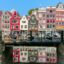 A row of narrow colourful houses with lots of windows reflected in a body of water with a bridge crossing over it and bikes parked along it
