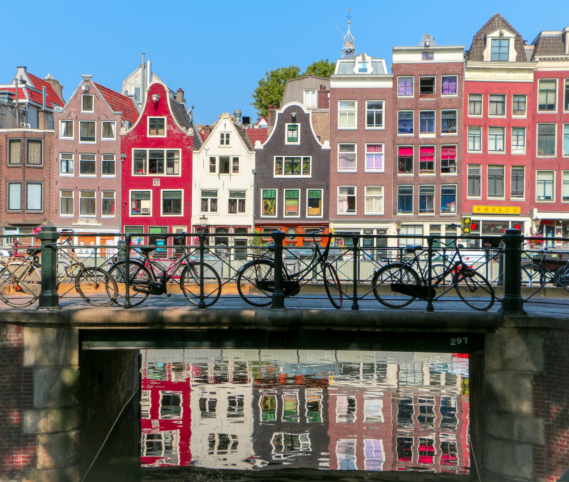 A row of narrow colourful houses with lots of windows reflected in a body of water with a bridge crossing over it and bikes parked along it