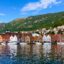 Colorful houses and small motor boats line the waterway of Bergen, backed by tree-covered mountains and a blue sky.