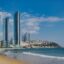 A view of Gwangalli Beach in Busan on a clear sunny day with three very tall skyscrapers along with other buildings set back from the shoreline in the distance.
