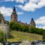 A view up a green tree-lined hill of the Westpomeranian building in Szczecin with its two notable towers, with a stone cathedral to the left.