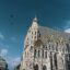 A view of Stephansdom cathedral in Vienna with its baroque and gothic features and a mosaic roof set against a blue sky with birds flying in view.
