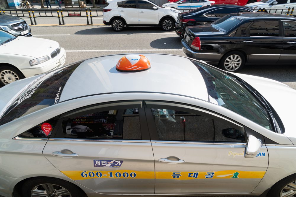 A white Busan taxi with an orange taxi light on the roof and a yellow strip across the doors with the company details displayed on it.