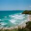 the colorful whitecaps upon aqua waters alongside the sandy and lush coast of Byron Bay under a blue sky