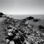 A black and white photo of a beach with pebbles and jagged rocks along the shoreline.