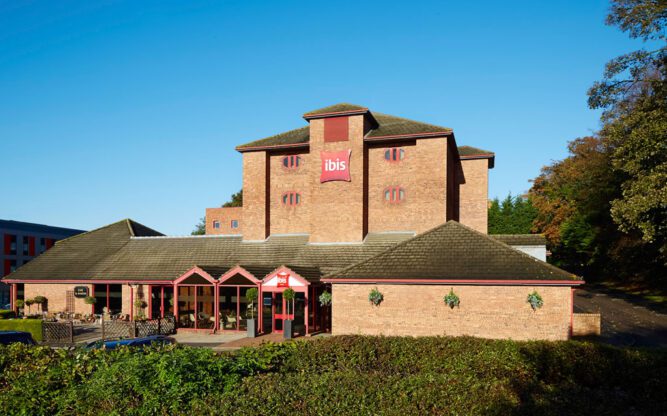 Exterior of ibis Styles at Luton Airport, with blue skies above and green grass surrounding the hotel.